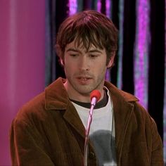 the young man is talking into a microphone while wearing a brown jacket and white t - shirt