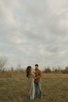 a man and woman standing in a grassy field under a cloudy sky with trees behind them