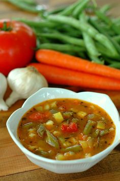 a white bowl filled with soup next to green beans, carrots and tomatoes on a cutting board