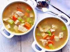 two bowls of chicken and vegetable soup on a white wooden table with silver spoons