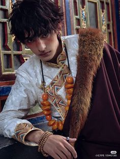 a young man wearing an elaborately decorated coat and beaded bracelets sits on a bench