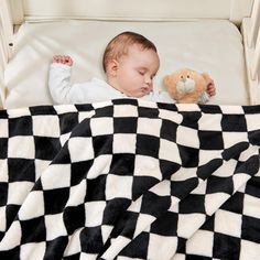 a baby sleeping in a crib with a teddy bear and checkerboard blanket