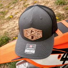 This customized hat is great for every dirt bike riding or motocross lover. The leather patch in front will come with your state letters on either side of the cross arrows and state map on the bottom. The top emblem will be the dirt bike or you can customize this by selecting another one from out options list. If you are looking for a different emblem please let us know and we will see if we can find it. This is a one size fits most, adjustable snap back Richardson original 112 trucker hat in gr Adjustable Cap For Biker Events, Adjustable Black Motorcycle Hat, Biker Cap For Outdoor Use, Biker Cap For Outdoor, Outdoor Biker Cap, Biker Style Cap For Outdoor, Custom Outdoor Baseball Cap With Flat Bill, Custom Snapback Baseball Cap For Outdoor, Dirt Bike Riding