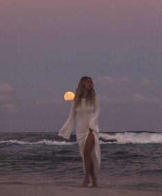 a woman standing on top of a beach next to the ocean at night with a full moon in the sky