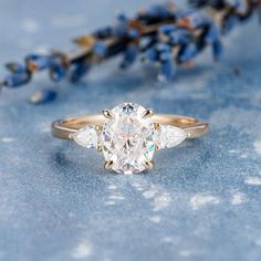 an oval cut diamond ring with three pear shaped diamonds on the side, sitting next to blue flowers