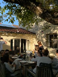 a group of people sitting around a table eating food under a tree in front of a house