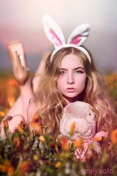 a woman laying in the grass holding a teddy bear with bunny ears on her head