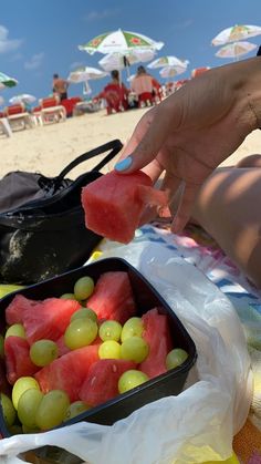 watermelon and grapes in a plastic container on the beach with people sitting under umbrellas