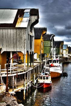 several boats are docked in the water next to some wooden buildings and cloudy skies above them