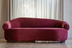 a red couch sitting on top of a hard wood floor next to a curtained window