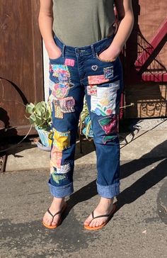 a woman standing in front of a wooden fence wearing jeans with patches and flowers on them