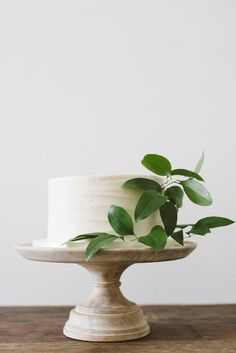 a white cake with green leaves on top sitting on a wooden table next to a potted plant