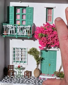 a hand is holding up a doll house with green shutters and pink flowers on the balcony