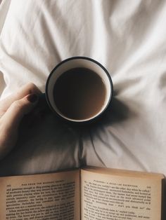 an open book with a cup of coffee in it on top of a bed next to a person's hand