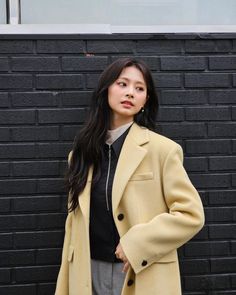 a woman standing in front of a black brick wall wearing a tan coat and grey pants