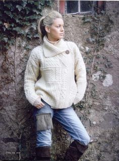 a woman leaning against a stone wall wearing a white sweater and jeans with a brown button