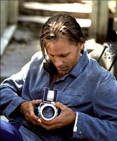 a man sitting on a bench holding an old camera and looking at his cell phone