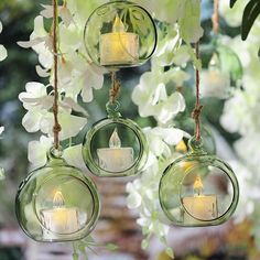 four glass hanging tea light holders with candles in them and white flowers on the side