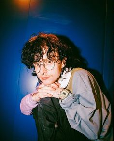 a young man with curly hair and glasses posing for the camera in front of a blue wall