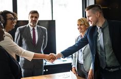 two business people shaking hands in front of a group of men and women at a desk