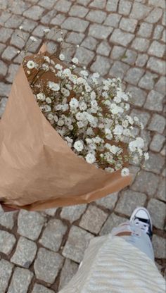 a person holding a brown paper bag with white flowers in it while standing on cobblestone pavement