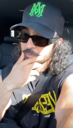 a man wearing sunglasses and a hat sitting in the back seat of a car with his hand on his chin