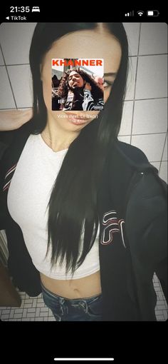 a woman with long black hair standing in front of a tile wall and wearing a white shirt