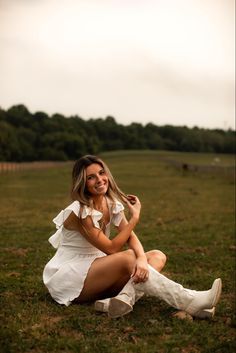 a woman sitting on the ground in a field with her legs crossed and wearing white boots