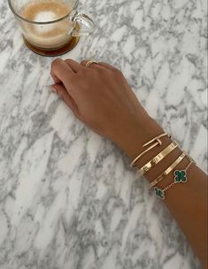 a woman's hand with four gold bracelets on it next to a cup of coffee