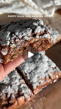 a hand is holding up some brownies with powdered sugar on top and the words puppy chow rice krispies treats below it