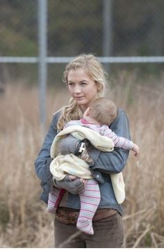 a woman holding a baby in her arms while standing next to a field with tall grass