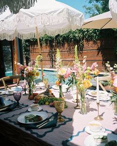 an outdoor dining table set with flowers and plates under umbrellas next to a swimming pool