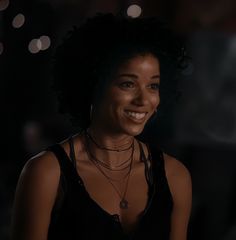 a woman with black hair smiling and wearing necklaces on her neck, in a dark room