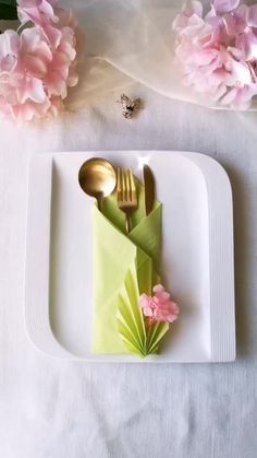 a white plate topped with a green napkin and two gold spoons next to pink flowers