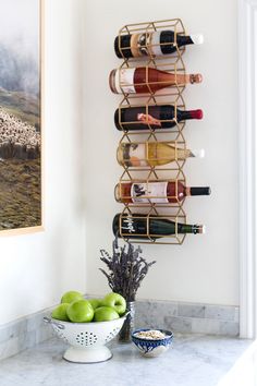 a wall mounted wine rack holding several bottles and two bowls filled with fruit on a marble countertop