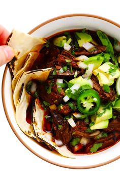 a hand holding a tortilla filled with meat and vegetables in a white bowl