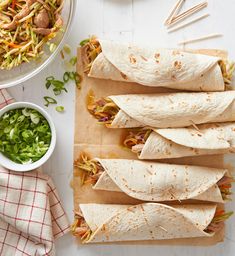 four burritos are lined up on a cutting board next to a bowl of salad and chopsticks