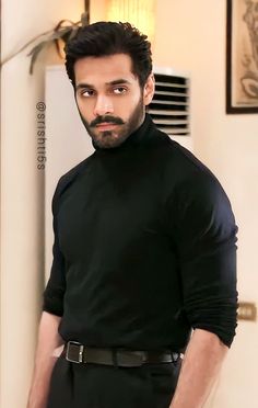 a man with a beard wearing a black shirt and pants standing in front of a wall