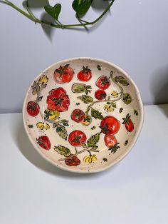 a bowl with tomatoes painted on it sitting next to a green plant in front of a white wall