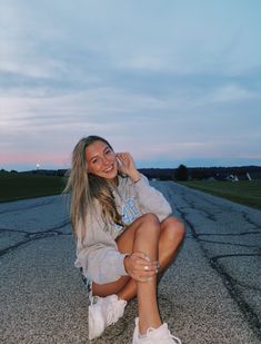 a woman sitting on the ground with her legs crossed and wearing white sneakers, smiling at the camera