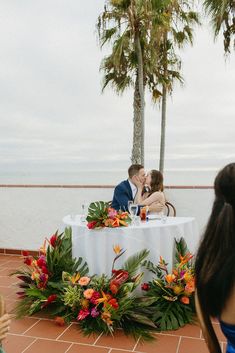 two people sitting at a table with flowers and greenery on it, surrounded by palm trees