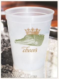 there is a plastic cup that has a crocodile on it and the words cheers written in gold