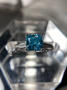 a fancy blue diamond ring sitting on top of a clear glass block with diamonds around it