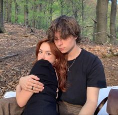 a man and woman sitting on a bench in the woods with their arms around each other