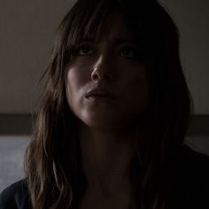 a woman with long brown hair and blue eyes stares into the distance while standing in a dimly lit room