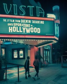 two people standing in front of a movie theater at night with the hollywood sign lit up