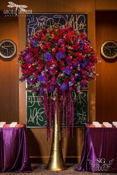 a tall vase filled with purple and red flowers next to two tables covered in purple tablecloths