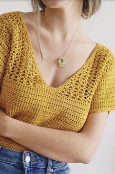 a woman with her arms crossed wearing a yellow sweater and necklace, standing in front of a white wall