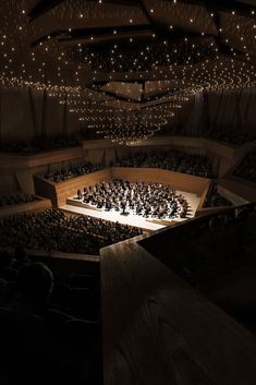 an auditorium filled with lots of people sitting on top of a wooden stage and lights hanging from the ceiling