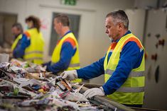 two men in yellow vests are sorting items at a recycling center with other workers